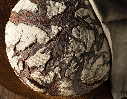Ein Holzofenbrot auf einem runden Holzbrett, auf der rechten Seite etwas abgedeckt mit einem Leinentuch